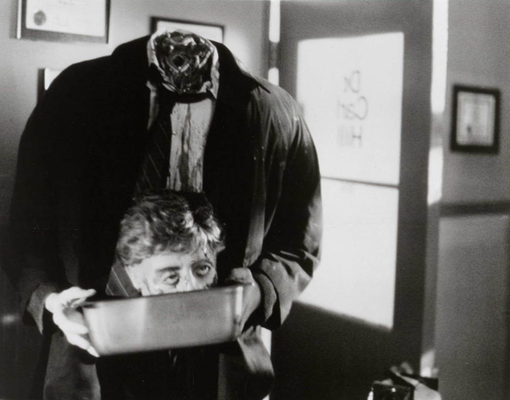 Film still from Re-Animator. A headless animated corpse carries its head in a metal bin.