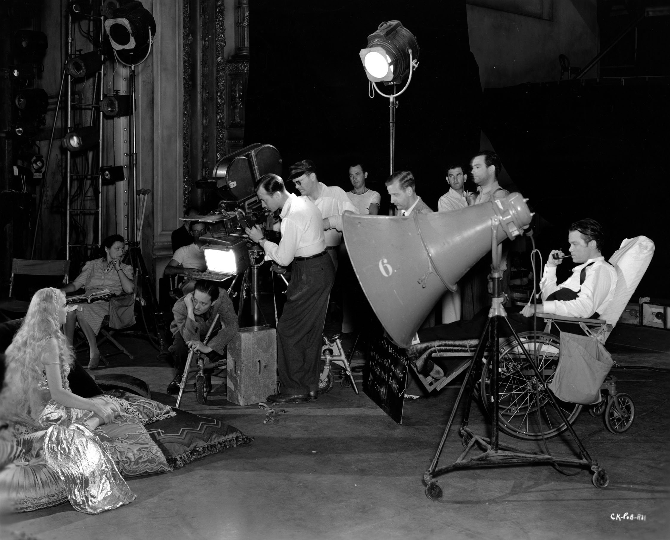 Production still. Original caption reads: "ORSON FILMS AN OPERA. Directing from a wheelchair while an injured ankle heals, Orson Welles, right, is shown here overseeing rehearsals of an opera scene for his initial RKO Radio production, “Citizen Kane.” Welles plays the title role in the picture, also produces and directs. Seated left is Dorothy Comingore, the young 'unknown' Welles chose for a leading role in the film. Crouched before the camera is Gregg Toland, director of photography, while the balance
