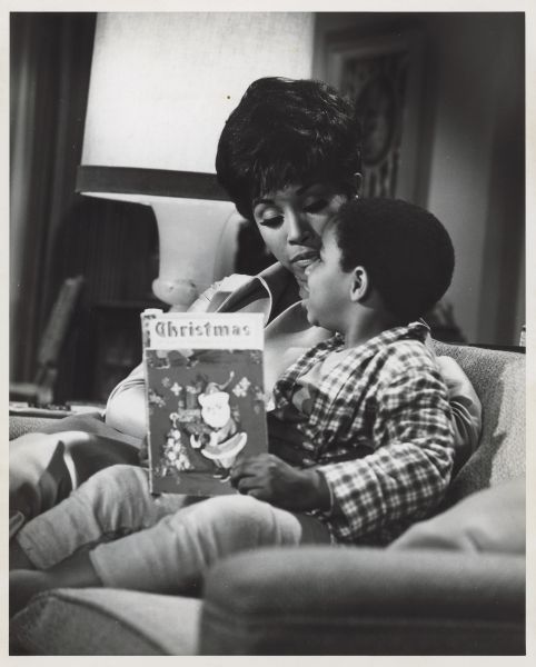 A black woman sitting on a couch with a black child, who is holding a Christmas book.