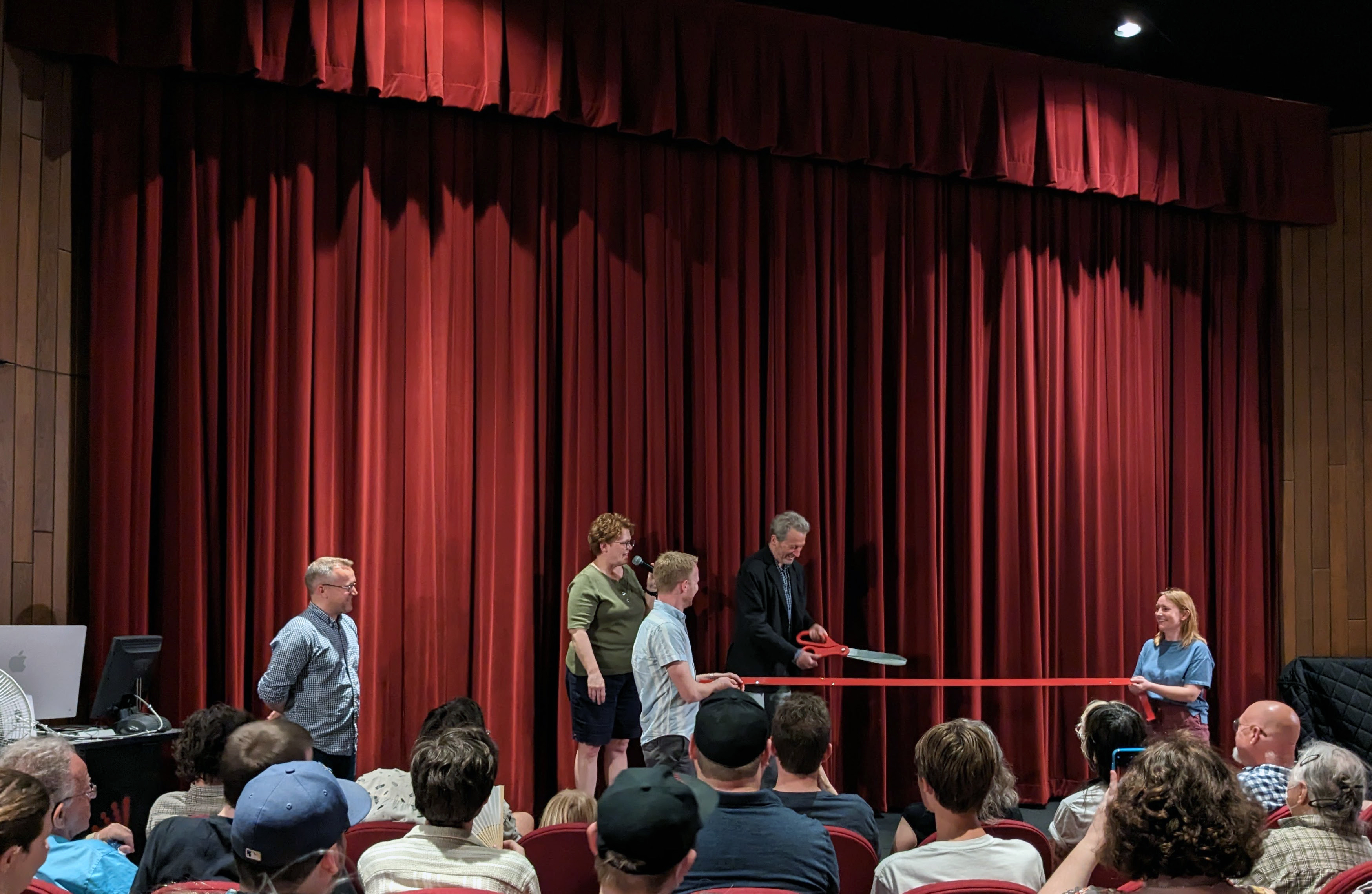 A large audience in a theater watches as Ken Kwapis prepares to cut a ribbon with a large pair of scissors