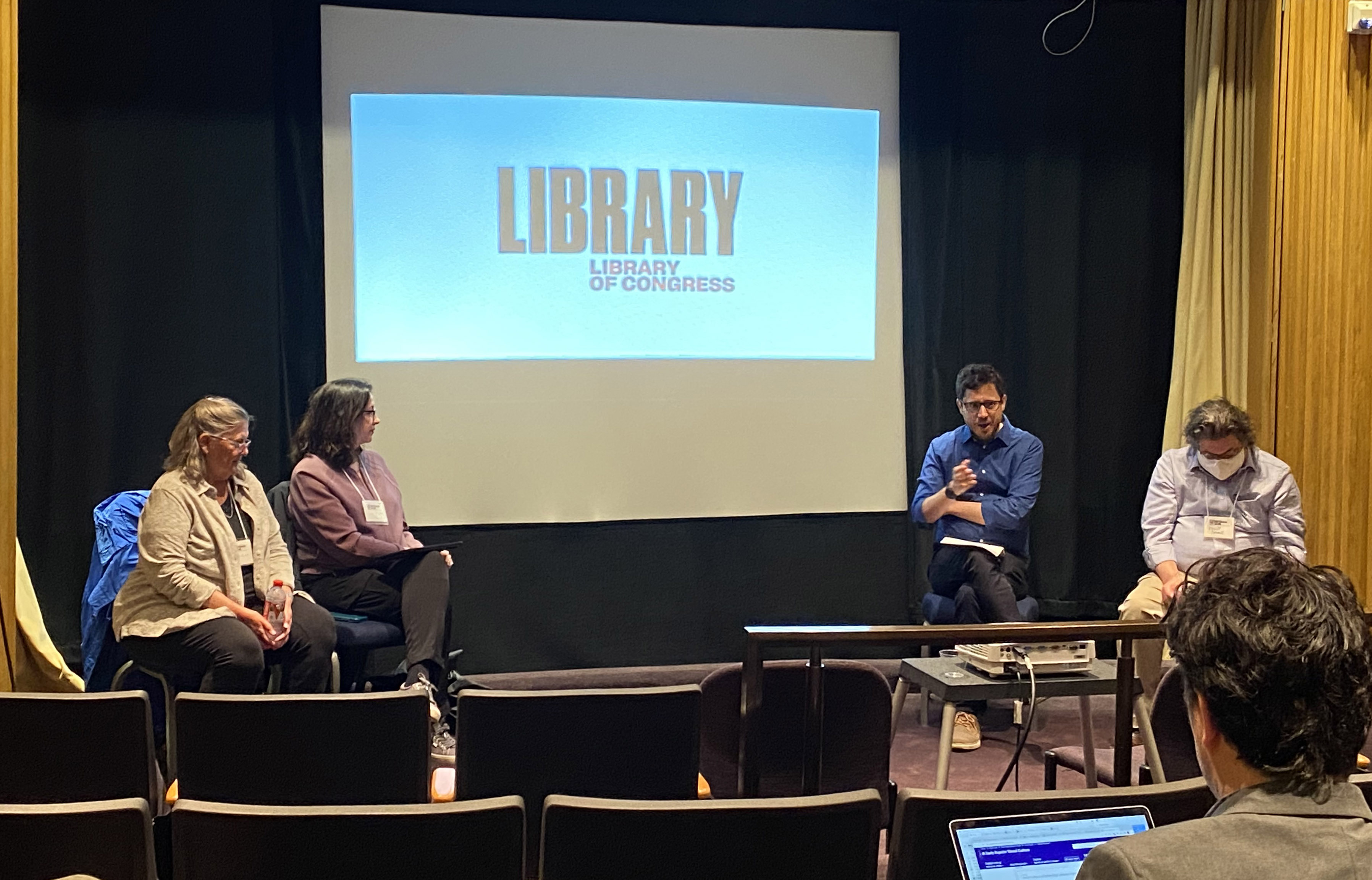 A photo of four people seated at the front of a theater for a panel discussion. The screen displays the logo for the Library of Congress