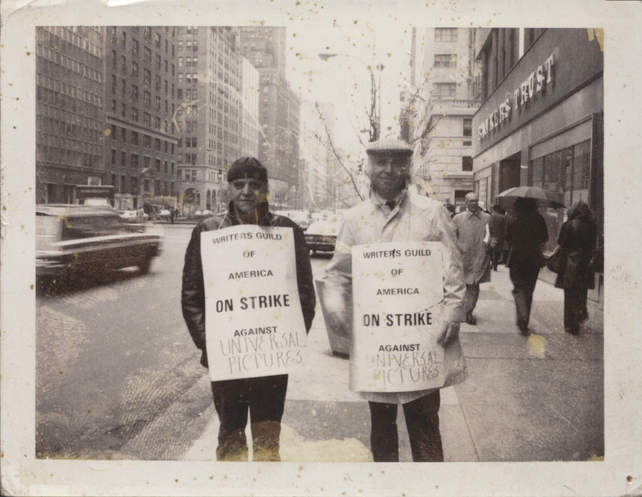 A scan of a black and white photo. Two men stand on the side of a busy street wearing signs which read "Writers Guild of America ON STRIKE Against Universal Pictures." The photograph has fingerprints on it and several small specks and smudges.