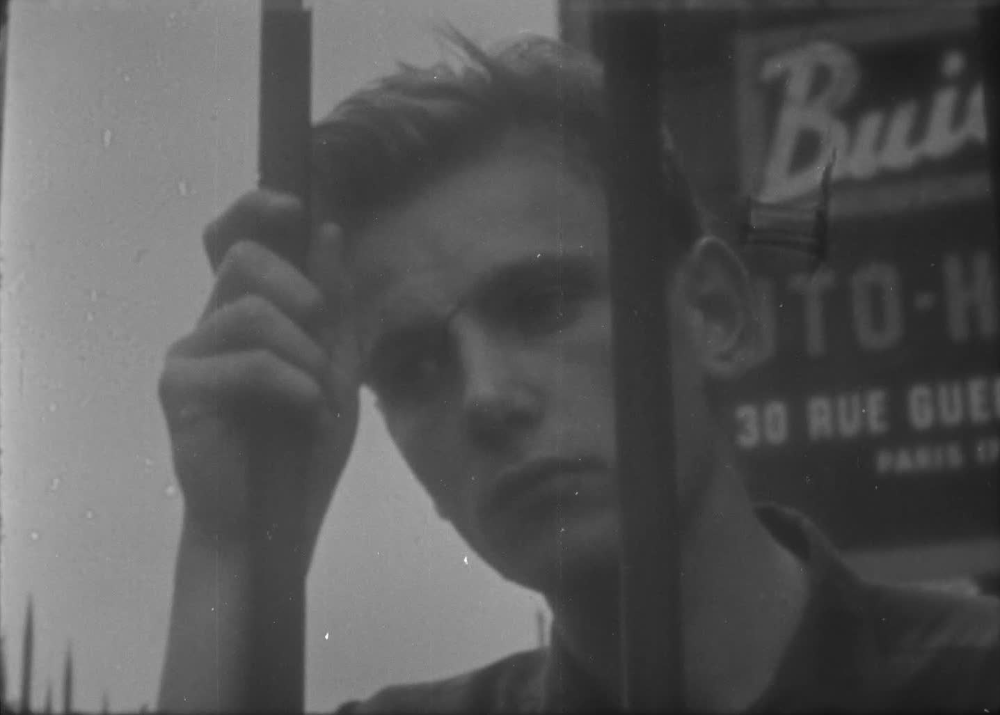 Black and white still frame from LAFCADIO. A man leans his head against a set of vertical bars, with his hand holding one of the bars.