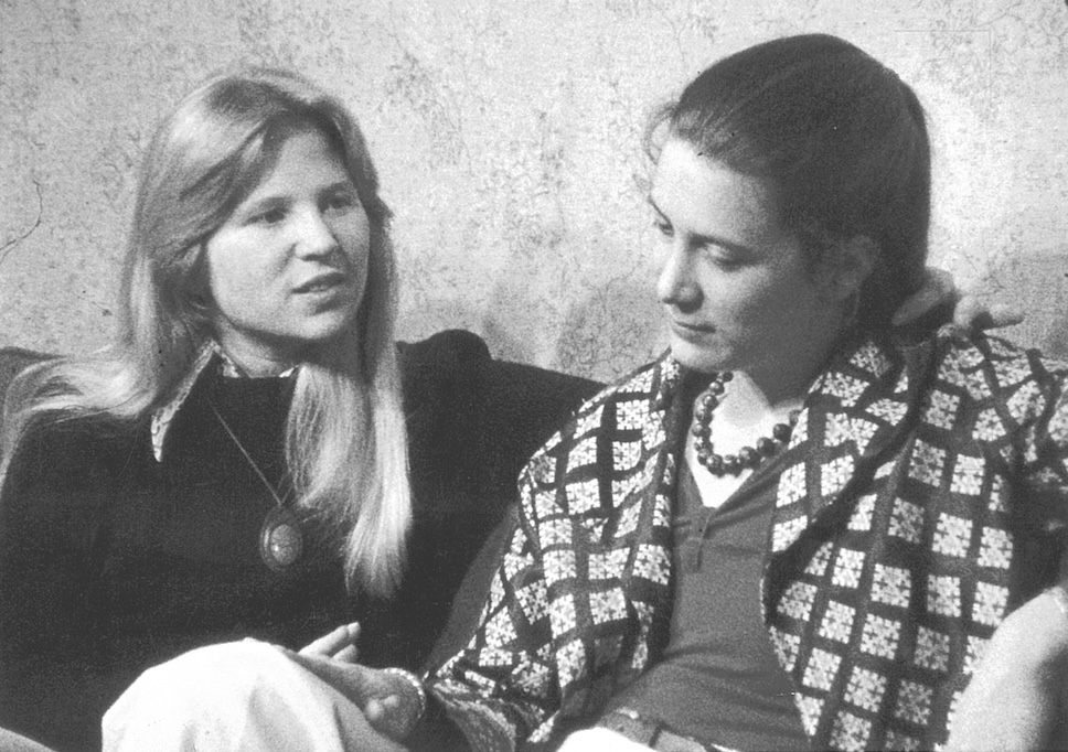 Two women sitting next to each other, with one draping an arm around the other's neck.