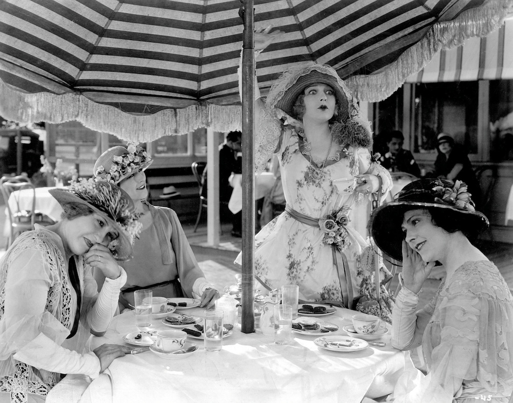 Four women wearing dresses and hats at a table, with one woman standing up.